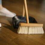 Close-up of a broom and dustpan with white sneakers indoors, representing housekeeping and cleanliness.